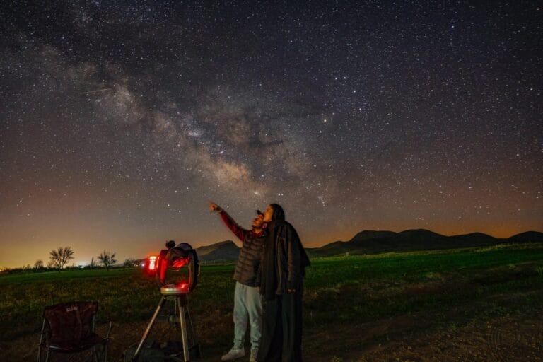 Two people stargazing in green meadows along with telescope