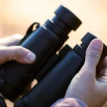 A person holding a pair of black compact binoculars in their hands